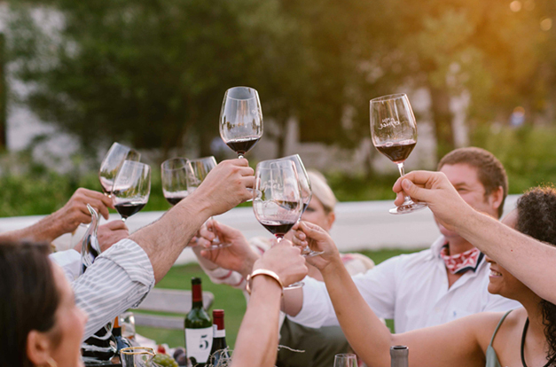 People celebrating outdoors at sunset, raising their red wine glasses for a toast