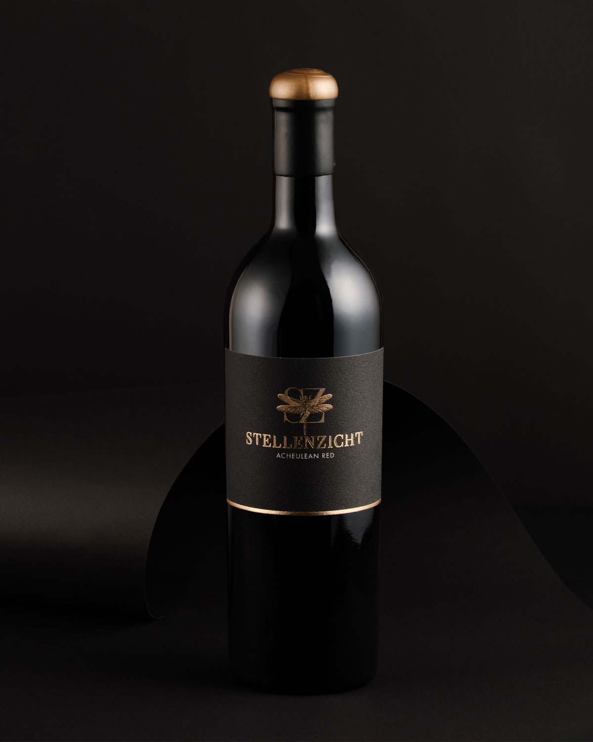 Eine dunkle Weinflasche mit einem schwarz-goldenen Etikett, das eine Libelle und den Text Stellenzicht Acheulean Red zeigt. Der Deckel der Flasche ist mit einer Goldfolie überzogen.