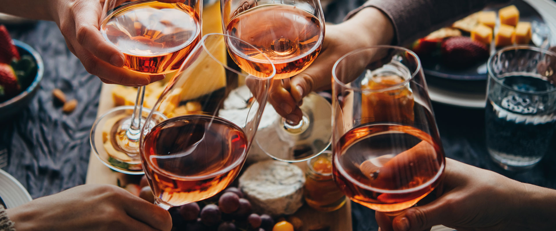 Friends toast with glasses of rosé wine, surrounded by cheese and fruit on a laid table