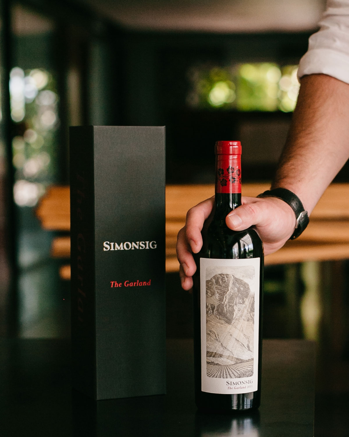 Eine Rotweinflasche mit rotem Verschluss und schwarzem Blumenmuster. Das Etikett trägt die Aufschrift SIMONSIG The Garland und zeigt eine detaillierte Illustration einer Berglandschaft mit Feldern im Vordergrund.