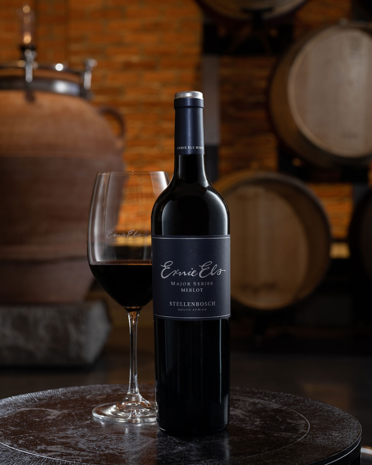 Eine Flasche Ernie Els Major Series Merlot und ein gefülltes Weinglas stehen auf einem runden Tisch mit großen Holzfässern und einer braunen Ziegelwand im Hintergrund.