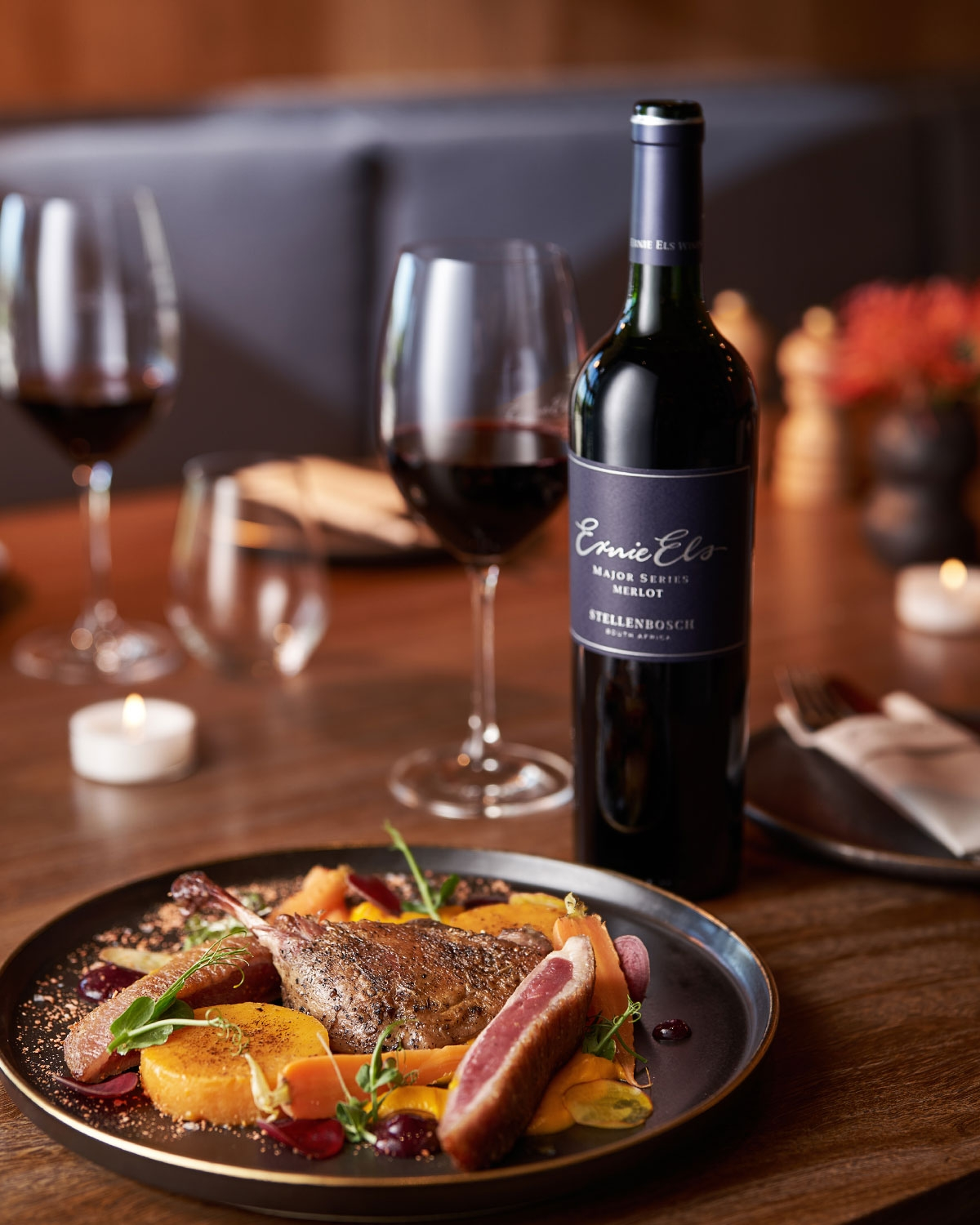 Ein Teller mit Gourmet-Entenbrust mit Gemüse und Soße wird auf einem Holztisch serviert, daneben eine Flasche Rotwein mit der Aufschrift Ernie Els Merlot und zwei gefüllte Weingläser, im Hintergrund Kerzen und gedeckte Tische.