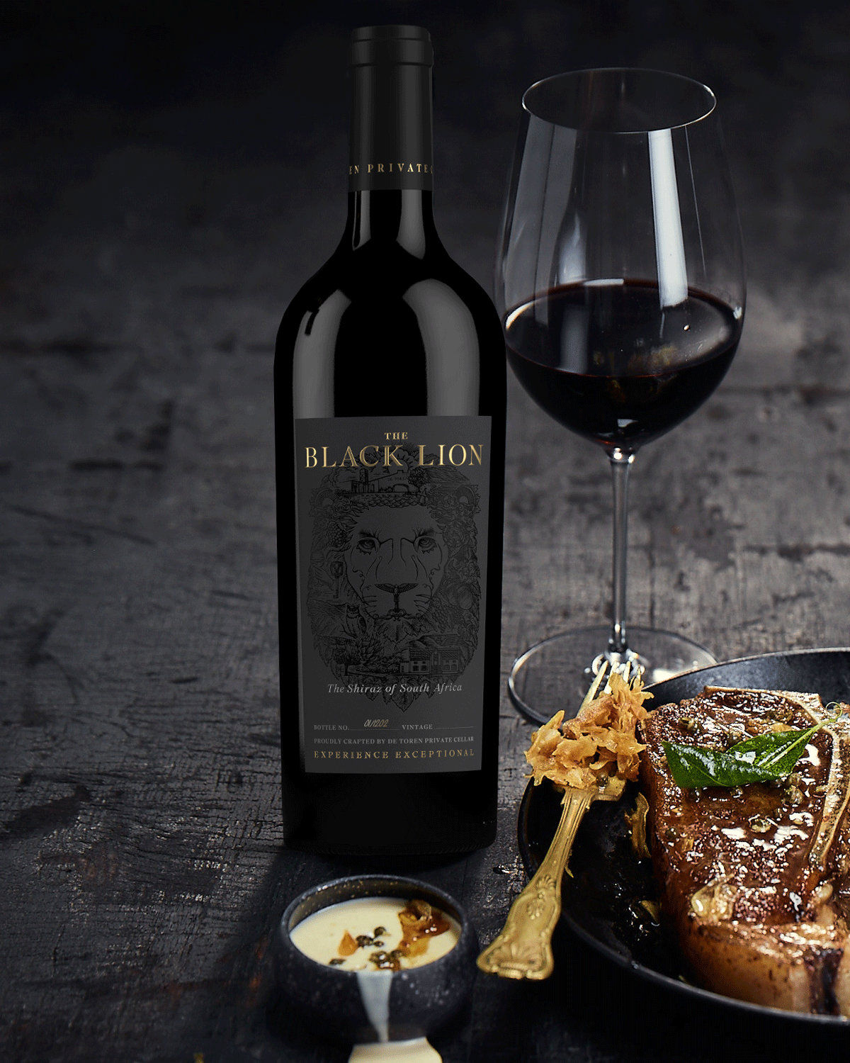 Eine Flasche des Rotweins The Black Lion steht neben einem Glas Rotwein und einem Teller mit einem gebratenen Steak, Soße und Beilage auf einer dunklen Oberfläche.